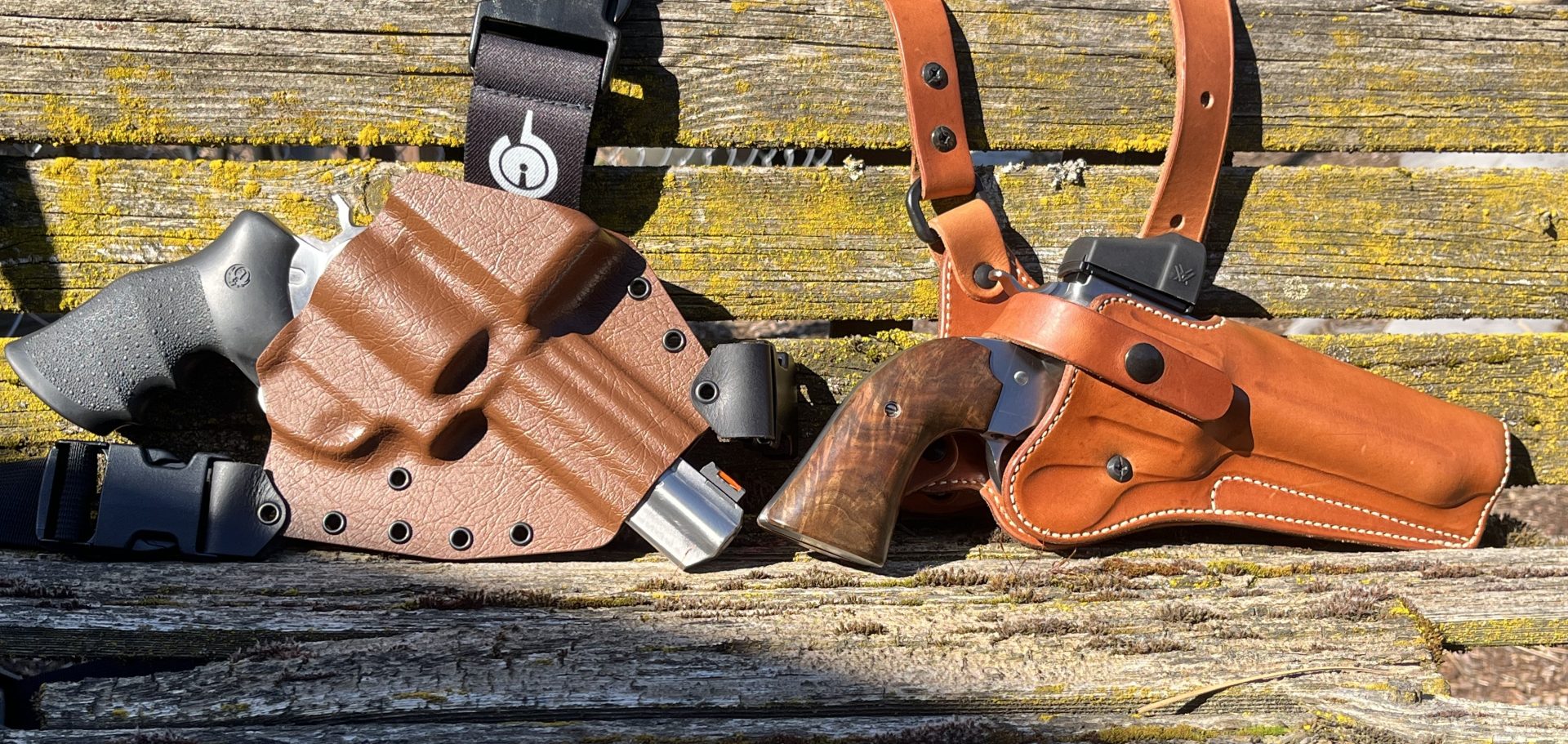 Two revolvers in leather chest holsters resting on weathered wooden boards outdoors.
